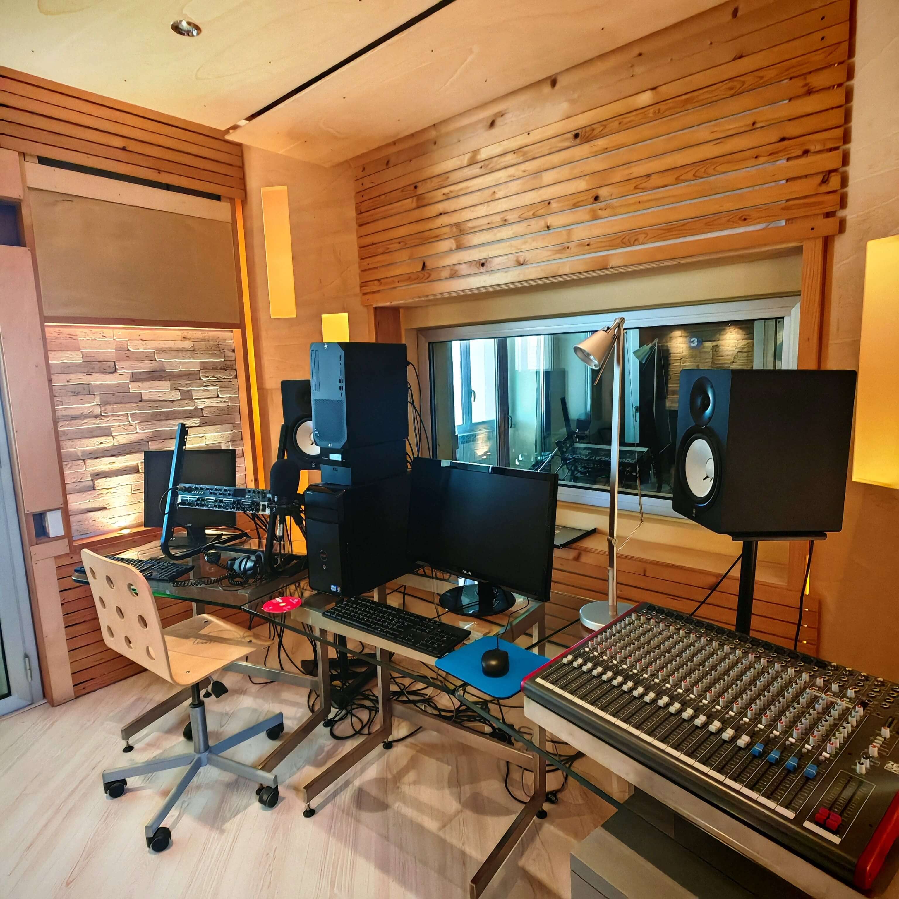 Postazione di regia audio con mixer, monitor, casse monitor e pareti trattate acusticamente con listelli di legno.